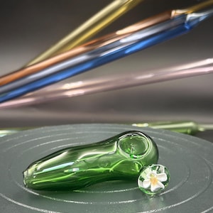 May include: A green glass pipe with a rounded bowl and a small, clear glass marble with a white flower inside. The background features several glass rods in gold, blue, and pink.