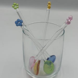 以下が含まれることがあります： 4本の装飾的なマドラーが入った透明なグラス。各マドラーは、緑、青、黄色、ピンクの異なる色の花型のトップが付いています。グラスには、カラフルなフルーツ型の装飾も含まれています。