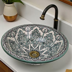 Könnte beinhalten: Ein ovales Keramikwaschbecken mit einem dekorativen türkis-weißen Muster. Das Waschbecken hat einen goldenen Abfluss und einen Wasserhahn aus dunkler Bronze. Das Waschbecken steht auf einer weißen Arbeitsplatte, unter einem Holzschrank.