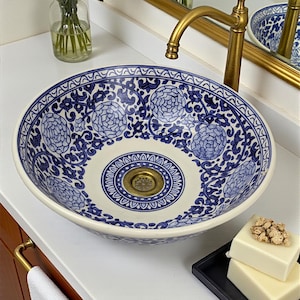 May include: A blue and white porcelain sink with a floral design. The sink is round and sits on a white countertop. A gold faucet and mirror are visible. Pink roses in a vase and soap are also present.