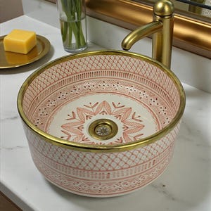 May include: A patterned ceramic bathroom sink with a gold rim, featuring a pink and white floral design. A gold tap and a vase of flowers are also visible, alongside a gold-framed mirror.