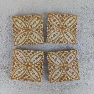 Könnte beinhalten: Vier quadratische Keramikuntersetzer mit floralem Design. Jeder Untersetzer hat einen cremefarbenen Hintergrund mit einem sich wiederholenden Muster aus vierblättrigen Blumen in einem warmen, goldbraunen Farbton. Die Untersetzer sind quadratisch angeordnet.