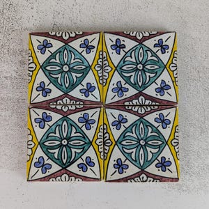 May include: Four square ceramic tiles with a repeating floral pattern. The design features teal, blue, yellow, and burgundy colors. The tiles are arranged in a square formation, showcasing the symmetrical pattern.