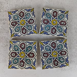 May include: Four square ceramic tiles with a repeating geometric pattern. The design features a central yellow starburst surrounded by teal, burgundy, and blue shapes, outlined in black. The tiles have a white background and yellow accents.