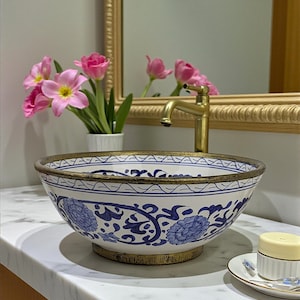 以下が含まれることがあります： 青と白の花柄と金色の縁が付いた装飾的な磁器製の洗面台。洗面台は大理石のカウンタートップにあり、金色の蛇口と小さな石鹸皿が付いています。白い花瓶に入ったピンクのチューリップが背景にあります。
