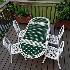 May include: Outdoor patio set with a green and white mosaic table and four white chairs. The table has a semi-circular end and is surrounded by a wooden deck with potted plants and a view of a lake.