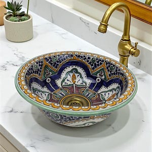May include: A decorative ceramic sink with a detailed, colourful pattern. The sink features a blue, white, green, and yellow design. It is mounted on a white countertop with a gold tap.