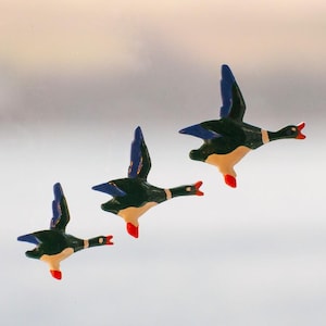 May include: Three duck figurines in flight, with black bodies, blue wings, and cream bellies. The beaks and feet are red. The background is a soft, blurred gradient of white and yellow.