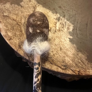 Op de afbeelding: Een trommelstok met een donkerbruine, ovaalvormige kop van viltmateriaal. De steel is van hout met een zwart-bruin patroon. Een band van wit materiaal bevindt zich tussen de kop en de steel. De achtergrond is een trommel.