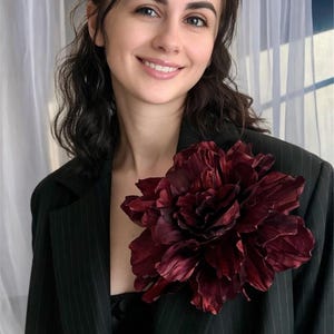 May include: A woman wearing a black pinstripe blazer with a large, burgundy flower brooch. The flower is the focal point, with detailed petals and a rich color. The blazer has a classic design.