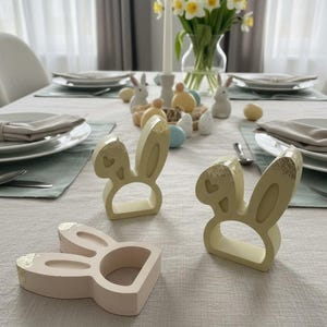 May include: Three bunny-shaped napkin rings in cream and pink with gold leaf detailing. The Easter table setting includes plates, napkins, and decorative eggs. A vase of daffodils and a candle complete the festive look.