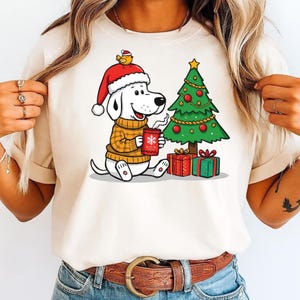 May include: A cream-coloured t-shirt with a cartoon dog wearing a Santa hat and a yellow sweater, holding a mug with a snowflake design. A decorated Christmas tree and wrapped gifts are also in the image. The dog has a small bird on its hat.