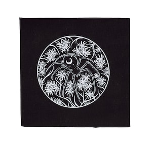 May include: A black fabric patch with a white circular design featuring a spider with a crescent moon on its back, surrounded by stylized flowers.