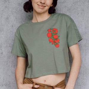 May include: Sage green crop top with a red tomato cluster graphic on the left chest. The shirt is paired with brown corduroy pants. The model is smiling in front of a gray wall.