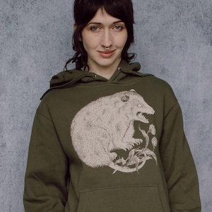 以下が含まれることがあります： オリーブグリーンのパーカー。ポッサムのイラストが描かれています。ポッサムは詳細なヴィンテージスタイルで描かれています。パーカーにはカンガルーポケットとドローストリングフードが付いています。イラストには花の要素が含まれています。