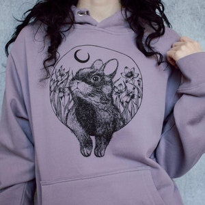 May include: A lavender colored hoodie with a black and white illustration of a rabbit in a circle with flowers and a crescent moon.