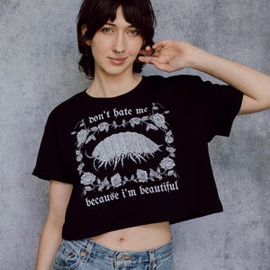 以下が含まれることがあります： ゴシック体で「don't hate me because I'm beautiful」と書かれた黒いクロップドトップ。ワラジムシのイラストと花のボーダーが特徴です。モデルはブルージーンズを着用しています。カジュアルなスタイルです。