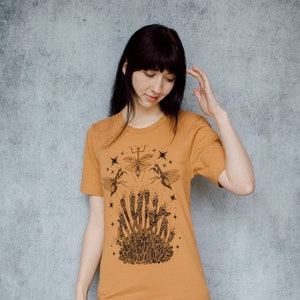 May include: A mustard yellow t-shirt with a black and white graphic print of a praying mantis, two bees, and stars. The graphic features the word "WHISPERS" in a stylized font.