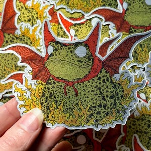 A die cut brushed silver metallic sticker of a full color toad in a devil costume with wings and flames.