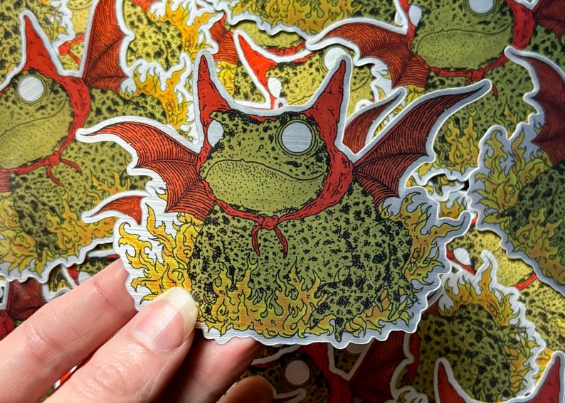 A die cut brushed silver metallic sticker of a full color toad in a devil costume with wings and flames.