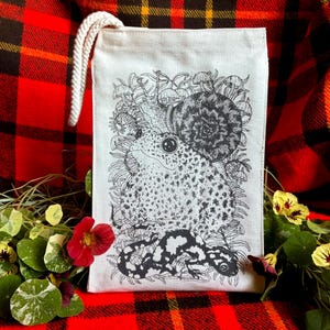 An off white natural canvas lunch bag with a rope handle and a deep charcoal grey hand screen printed design. The hand drawn natural illustration on the bag features a toad, a snail, a spotted salamander, ferns, and small mushrooms.