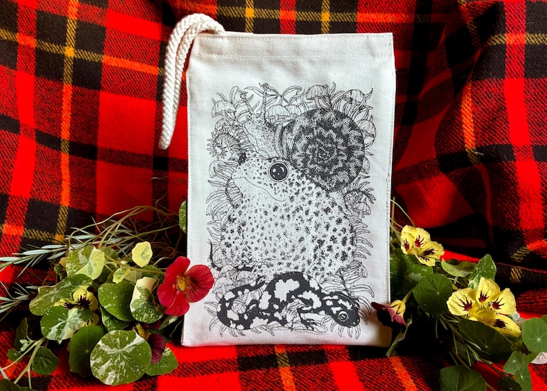 An off white natural canvas lunch bag with a rope handle and a deep charcoal grey hand screen printed design. The hand drawn natural illustration on the bag features a toad, a snail, a spotted salamander, ferns, and small mushrooms.