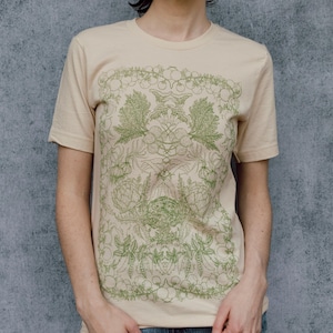 May include: A beige t-shirt with a green and white floral design. The design features a symmetrical pattern of leaves and flowers.