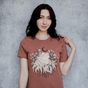 May include: A rust-colored t-shirt featuring a detailed graphic of a toad surrounded by flowers, leaves, and roots, with a crescent moon above. The design is in shades of cream and brown. The shirt has short sleeves and a crew neck.