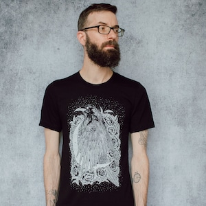 A unisex black t-shirt with a white hand screen printed design. The detailed hand drawn natural illustration is of a raven against a large crescent moon and peony flowers over a starry sky.
