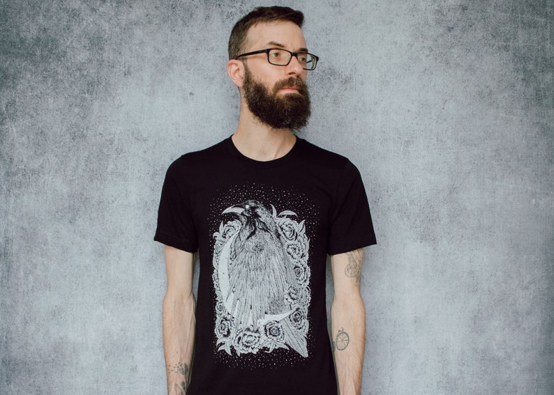A unisex black t-shirt with a white hand screen printed design. The detailed hand drawn natural illustration is of a raven against a large crescent moon and peony flowers over a starry sky.