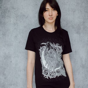 A unisex black t-shirt with a white hand screen printed design. The detailed hand drawn natural illustration is of a raven against a large crescent moon and peony flowers over a starry sky.