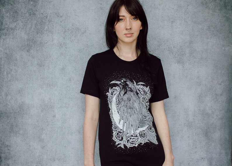 A unisex black t-shirt with a white hand screen printed design. The detailed hand drawn natural illustration is of a raven against a large crescent moon and peony flowers over a starry sky.