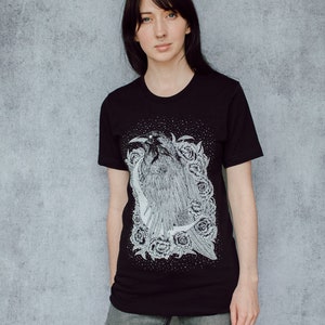 A unisex black t-shirt with a white hand screen printed design. The detailed hand drawn natural illustration is of a raven against a large crescent moon and peony flowers over a starry sky.