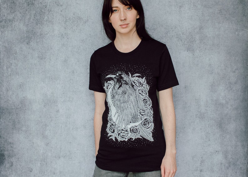A unisex black t-shirt with a white hand screen printed design. The detailed hand drawn natural illustration is of a raven against a large crescent moon and peony flowers over a starry sky.