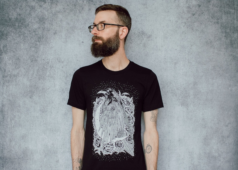 A unisex black t-shirt with a white hand screen printed design. The detailed hand drawn natural illustration is of a raven against a large crescent moon and peony flowers over a starry sky.