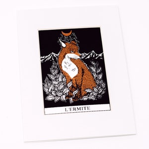 May include: A black and white illustration of a fox sitting in a forest with mountains in the background. The fox is orange and white and has a crescent moon above its head. The text "L'Ermite" is printed below the image.