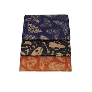 A set of three kraft cardstock pocket notebooks with natural illustration patters on the outside. One is print in blue ink with moths, one is black ink with skulls, and the third is red ink with various mushrooms and fungi.