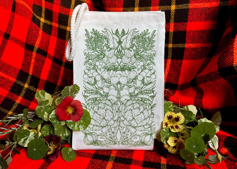 A natural colored canvas lunchbag featuring hand drawn art of various vegetables including artichokes, radishes, tomatoes, peas, chanterelle mushrooms, and garlic. The design is screen printed in warm green pink on the front of the bag.