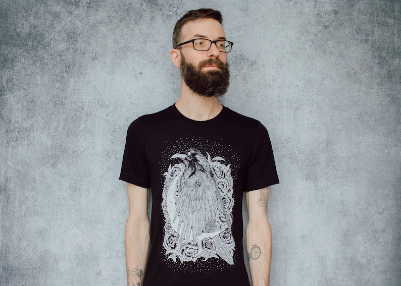 A unisex black t-shirt with a white hand screen printed design. The detailed hand drawn natural illustration is of a raven against a large crescent moon and peony flowers over a starry sky.