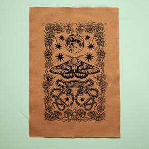 以下が含まれることがあります： 茶色の布地に、黒と白の蛾、満月、星、蛇のイラストが描かれたプリント。デザインは、花のつるで囲まれた枠で囲まれています。