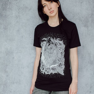 A unisex black t-shirt with a white hand screen printed design. The detailed hand drawn natural illustration is of a raven against a large crescent moon and peony flowers over a starry sky.