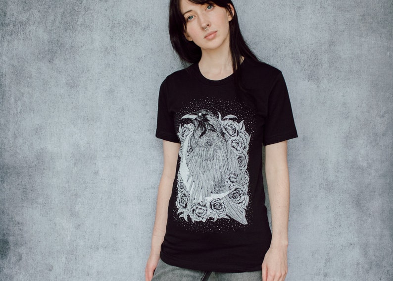 A unisex black t-shirt with a white hand screen printed design. The detailed hand drawn natural illustration is of a raven against a large crescent moon and peony flowers over a starry sky.
