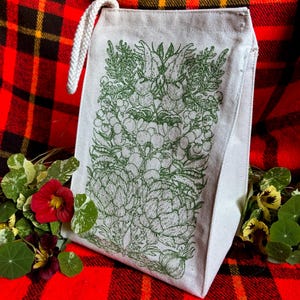 A natural colored canvas lunchbag featuring hand drawn art of various vegetables including artichokes, radishes, tomatoes, peas, chanterelle mushrooms, and garlic. The design is screen printed in warm green pink on the front of the bag.