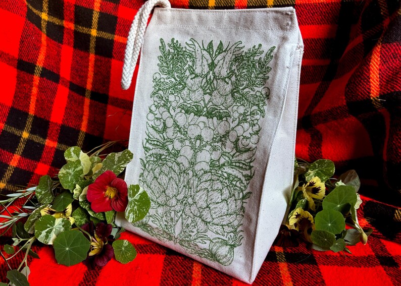 A natural colored canvas lunchbag featuring hand drawn art of various vegetables including artichokes, radishes, tomatoes, peas, chanterelle mushrooms, and garlic. The design is screen printed in warm green pink on the front of the bag.
