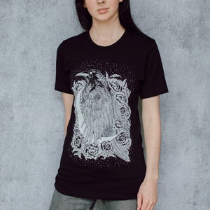 A unisex black t-shirt with a white hand screen printed design. The detailed hand drawn natural illustration is of a raven against a large crescent moon and peony flowers over a starry sky.
