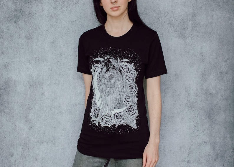 A unisex black t-shirt with a white hand screen printed design. The detailed hand drawn natural illustration is of a raven against a large crescent moon and peony flowers over a starry sky.