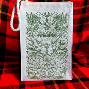 A natural colored canvas lunchbag featuring hand drawn art of various vegetables including artichokes, radishes, tomatoes, peas, chanterelle mushrooms, and garlic. The design is screen printed in warm green pink on the front of the bag.