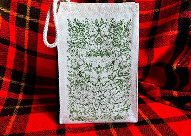 A natural colored canvas lunchbag featuring hand drawn art of various vegetables including artichokes, radishes, tomatoes, peas, chanterelle mushrooms, and garlic. The design is screen printed in warm green pink on the front of the bag.