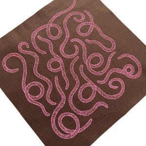 May include: A brown square textile with a pink, abstract embroidered design. The design features a series of interconnected, swirling lines and loops, creating a unique pattern. The textile has a textured appearance.