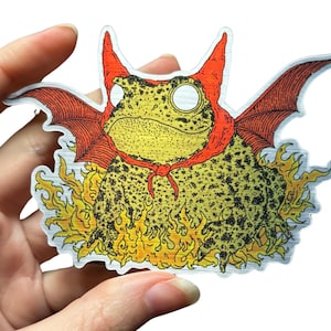 A die cut brushed silver metallic sticker of a full color toad in a devil costume with wings and flames.